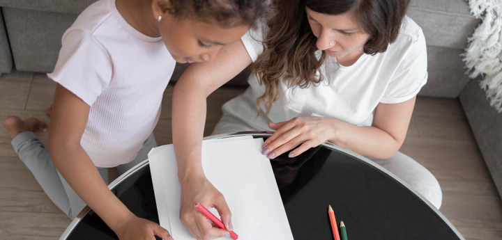 Mum and daughter sitting on floor in front of coffee table, with mum drawing on a piece of paper whilst the girl is looking over her shoulder at it