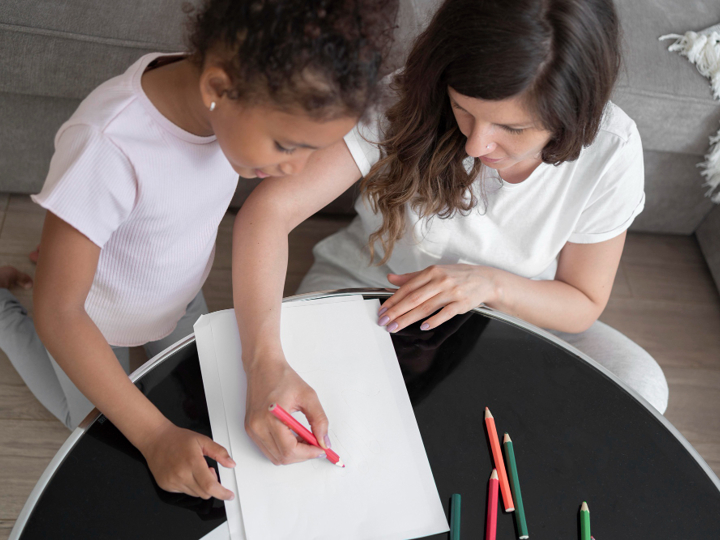 Mum and daughter sitting on floor in front of coffee table, with mum drawing on a piece of paper whilst the girl is looking over her shoulder at it