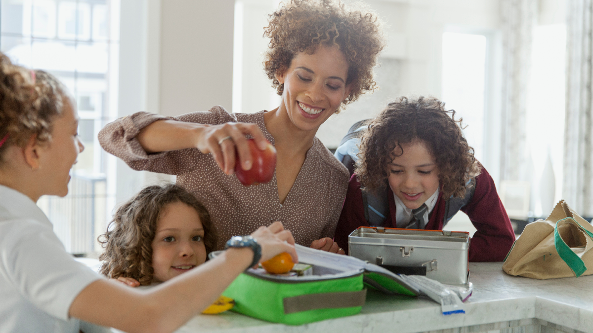 Woman standing in a kitchen surrounded by 3 children whilst she packs an apple into a lunchbox.