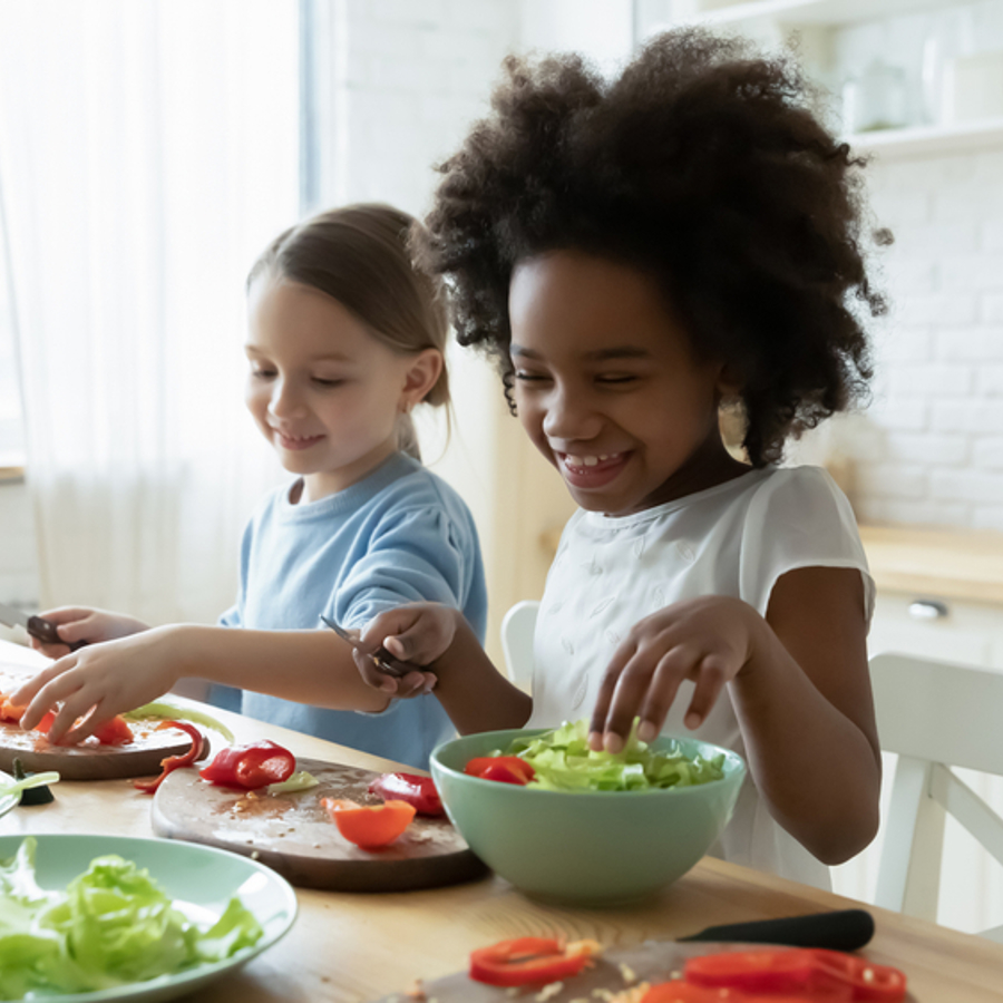 Two young girls sitting at kitchen table smiling and cutting up salad.