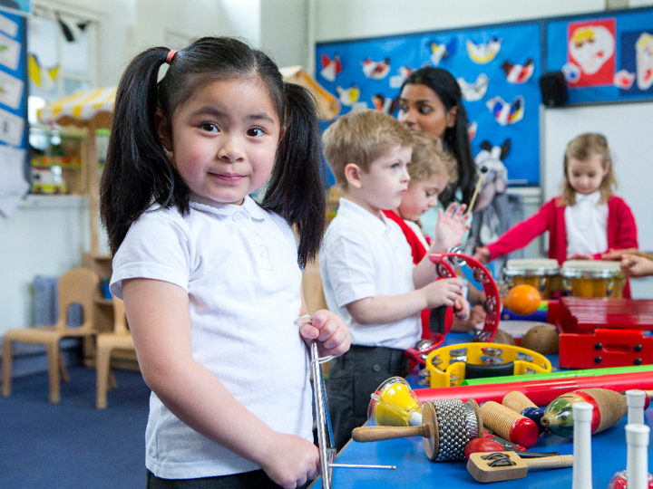 Primary school children in school uniform standing around a table playing with different musical instruments. A young girl in the foreground is staring directly into the camera.