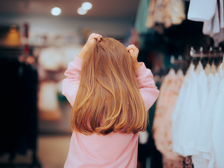 back of a child's head in a clothes shop. She's holding her hands to her head in a stressed gesture