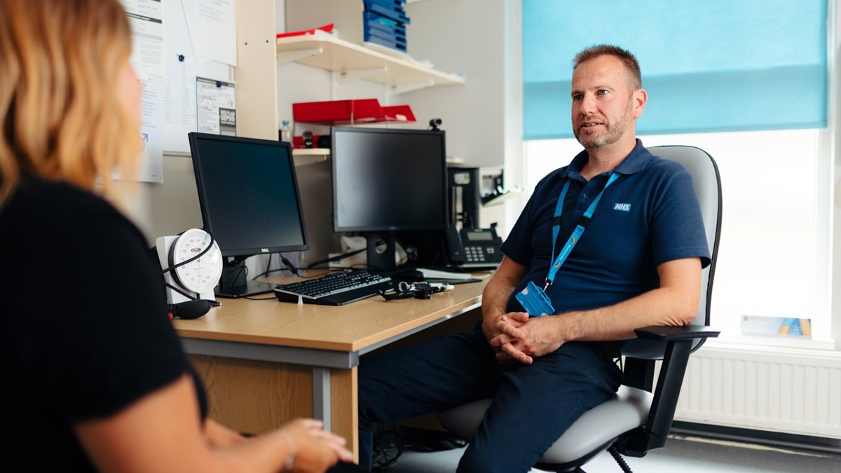 GP sitting at their desk, looking and speaking to a patient. The patient is in the foreground and out of focus on the left.