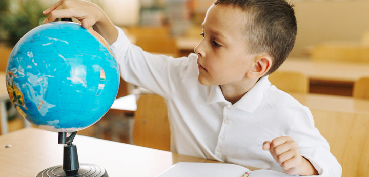 School boy in classroom sitting behind a desk with a notebook in front of him. He is turning a globe that is sitting on his desk