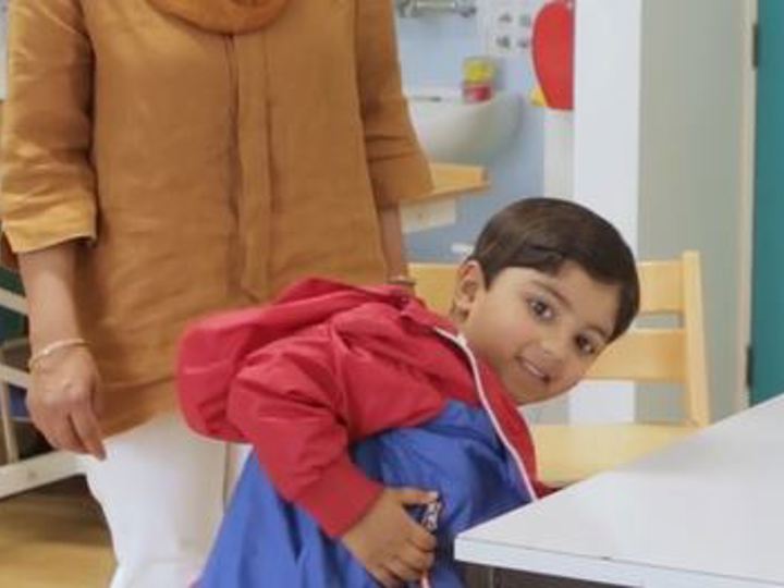 Child putting on a coat in a classroom with an adult in the background. The child has a cheeky smile.