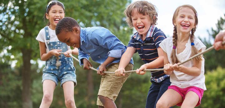 Four children pulling a rope in a game of tug of war whilst laughing and smiling outside in a park.