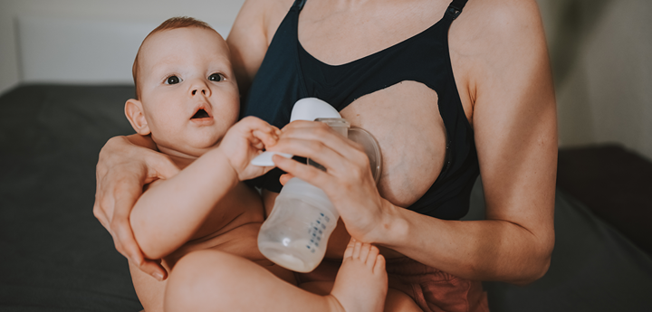 Woman sitting on sofa whilst expressing milk with a hand pump. Her other arm is holding her baby on her lap.