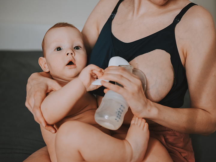 Woman sitting on sofa whilst expressing milk with a hand pump. Her other arm is holding her baby on her lap.