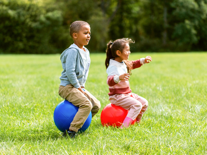 2 children outdoors on space hoppers jumping across a park and smiling