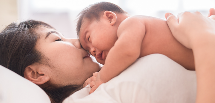 Woman lying on her back on a bed with a young naked baby lying face down on her stomach and chest.