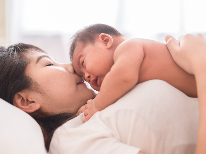 Woman lying on her back on a bed with a young naked baby lying face down on her stomach and chest.