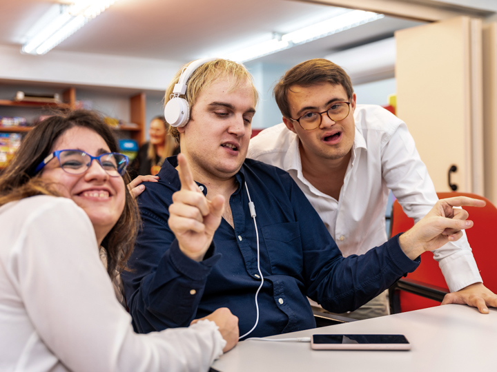 2 disabled men and a disabled woman sitting at a table in a community centre. The woman is on the left, smiling and hugging the man in the centre. The man in the centre is wearing headphones and is dancing. The man on the right is leaning forward and smiling at the camera.