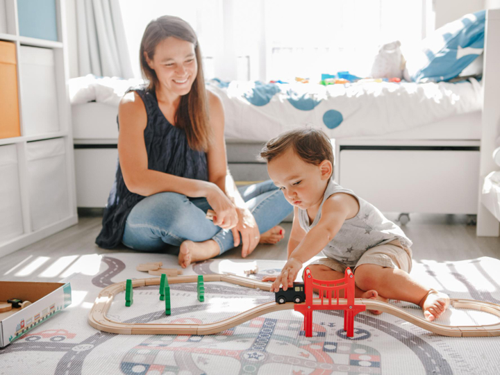 Boy toddler sitting on bedroom floor playing with train track. His mum is sitting next to him cross-legged on the floor watching him and smiling.