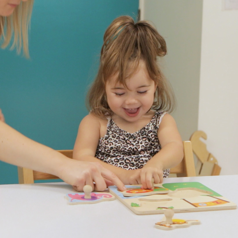 Young girl playing with a puzzle and smiling whilst an adult is pointing at a puzzle piece