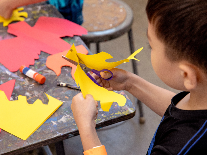 Little boy cutting shapes into yellow paper background is a black table with paint splattered over it with a gluestick and red, orange and yellow paper on