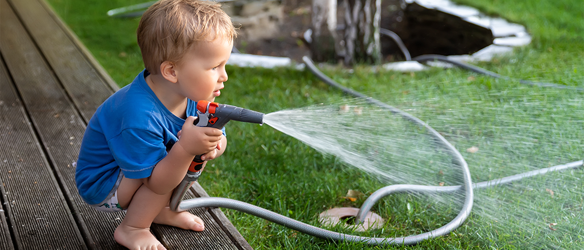 Child squatting using a garden hose to spray grass with water.