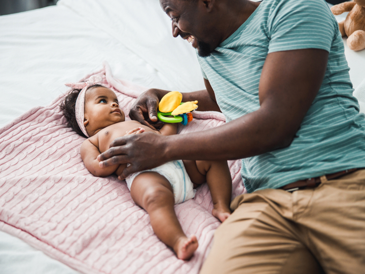 Young baby lying on a pink blanket on a bed looking at their smiling dad who is holding a toy.