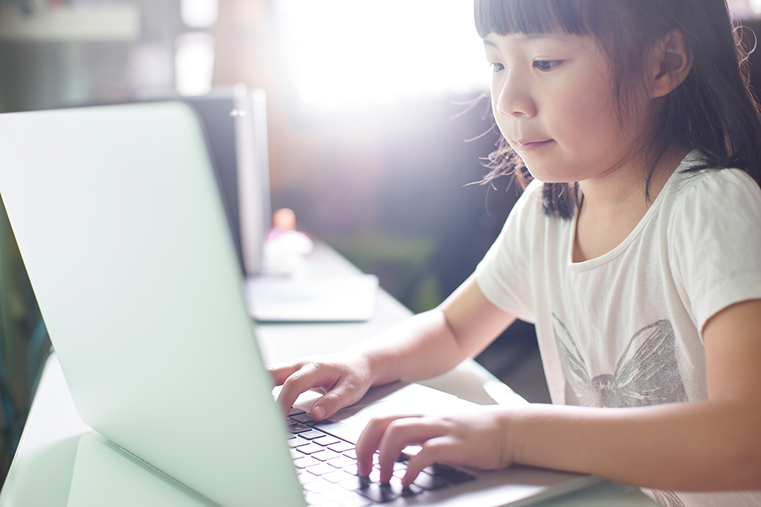 Young girl sitting at a table pressing keys on a laptop.