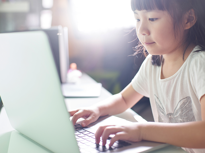 Young girl sitting at a table pressing keys on a laptop.