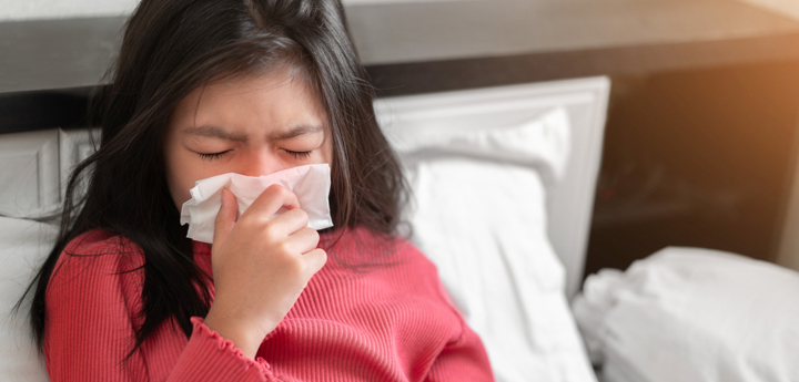 Girl sitting up in bed sneezing into a tissue.
