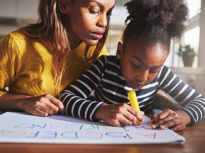 Young girl writing in coloured pens on a large sheet of paper. An adult woman is sitting behind the girl leaning over her shoulder and looking at what the girl is writing.