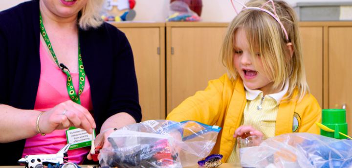 Girl in yellow cardigan and pink wire cat ears. Girl is playing with bags of plastic toys on a table. Sitting next to her is an adult helping.