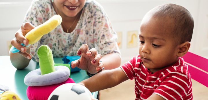 Toddler sitting at a child's table holding a football. A smiling adult is also sitting at the table and playing with the other soft toys on the table. 