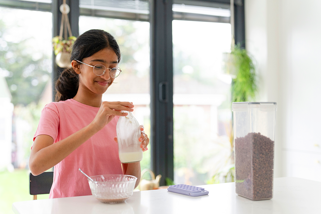 Teenage girl wearing glasses in kitchen holding a milk bottle over a bowl of cereal.