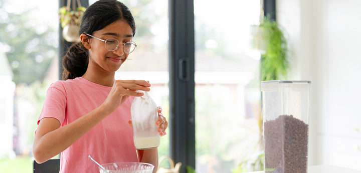 Teenage girl wearing glasses in kitchen holding a milk bottle over a bowl of cereal.