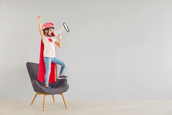 child standing on chair in a superhero costume wearing a helmet and cape holding a megaphone