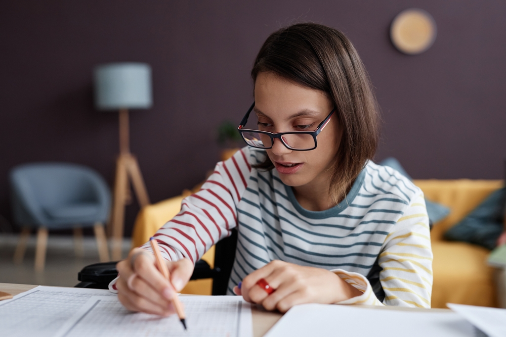Young adult writing in a lined booklet using a pen at table in their living room