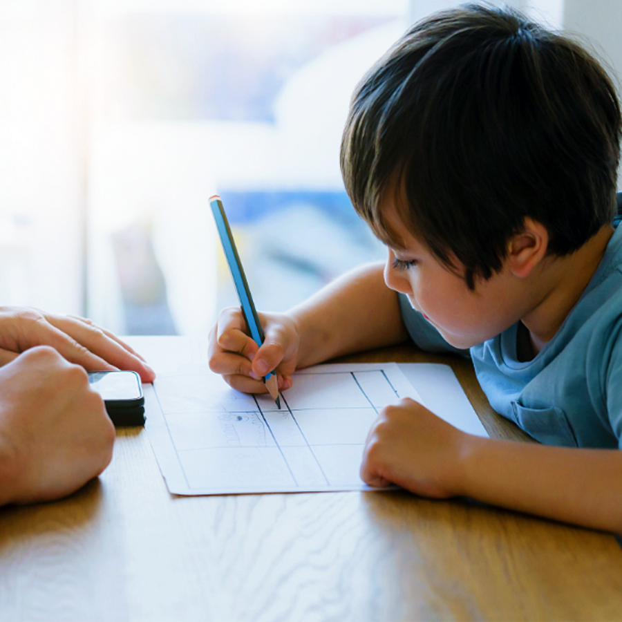 Young boy sitting at a table writing on a piece of paper with an adult sitting opposite of them.