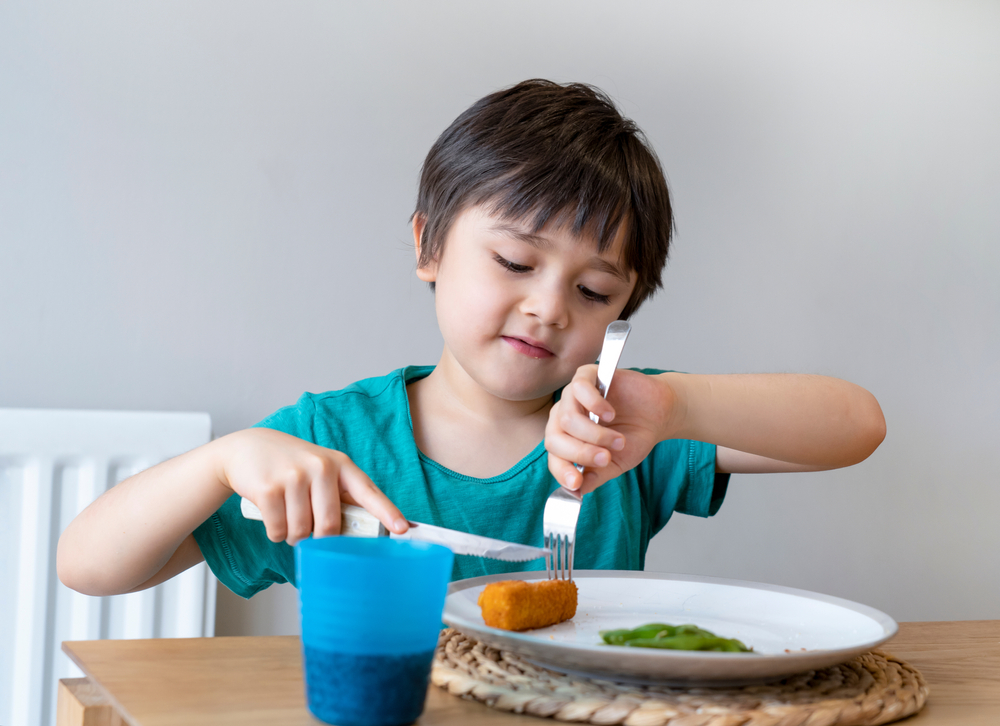 Child sitting at a table. They are cutting a fish finger using a knife and fork.