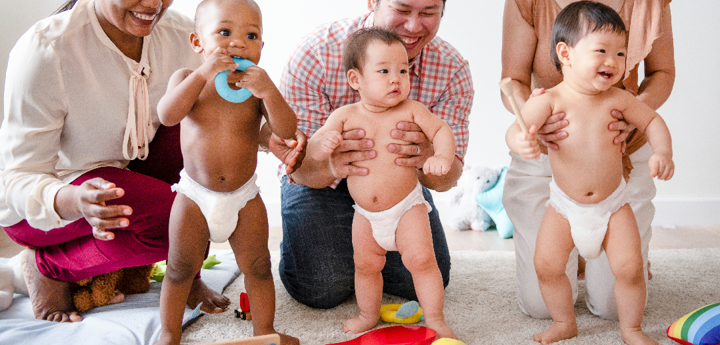 3 adults kneeling on the ground. 2 of the adults are holding their babies in a standing position. The other baby is standing independently holding a toy to their mouth in front of their smiling mother.