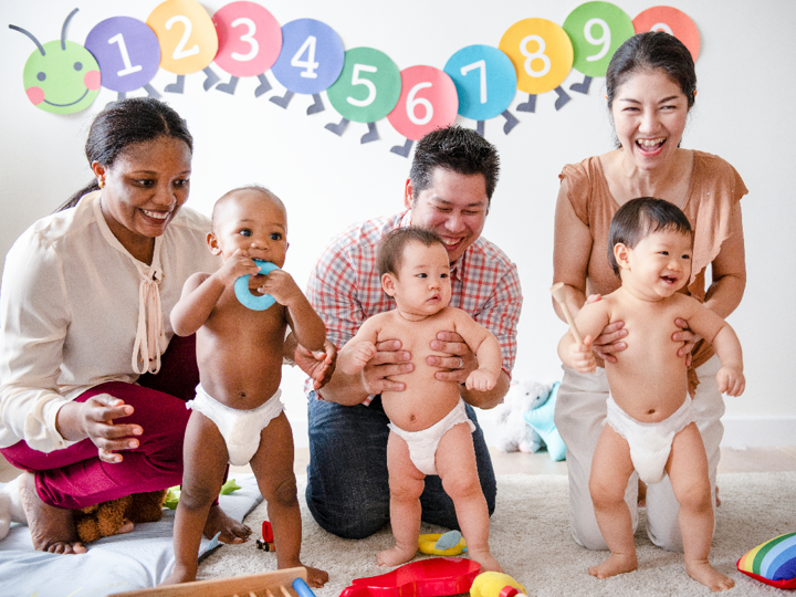 3 adults kneeling on the ground. 2 of the adults are holding their babies in a standing position. The other baby is standing independently holding a toy to their mouth in front of their smiling mother.