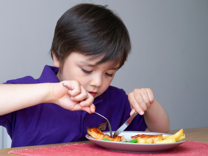 Boy sitting at table and using knife and fork to eat their food off their plate.