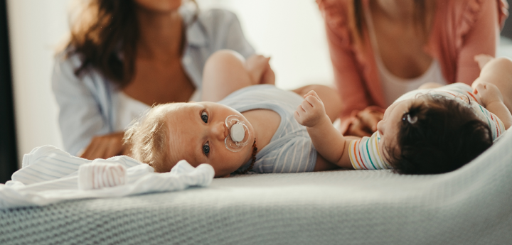 Two women kneeling down in front of their babies. The two babies are lying on their back. One baby is looking at the other, whilst the other baby is looking at the camera.