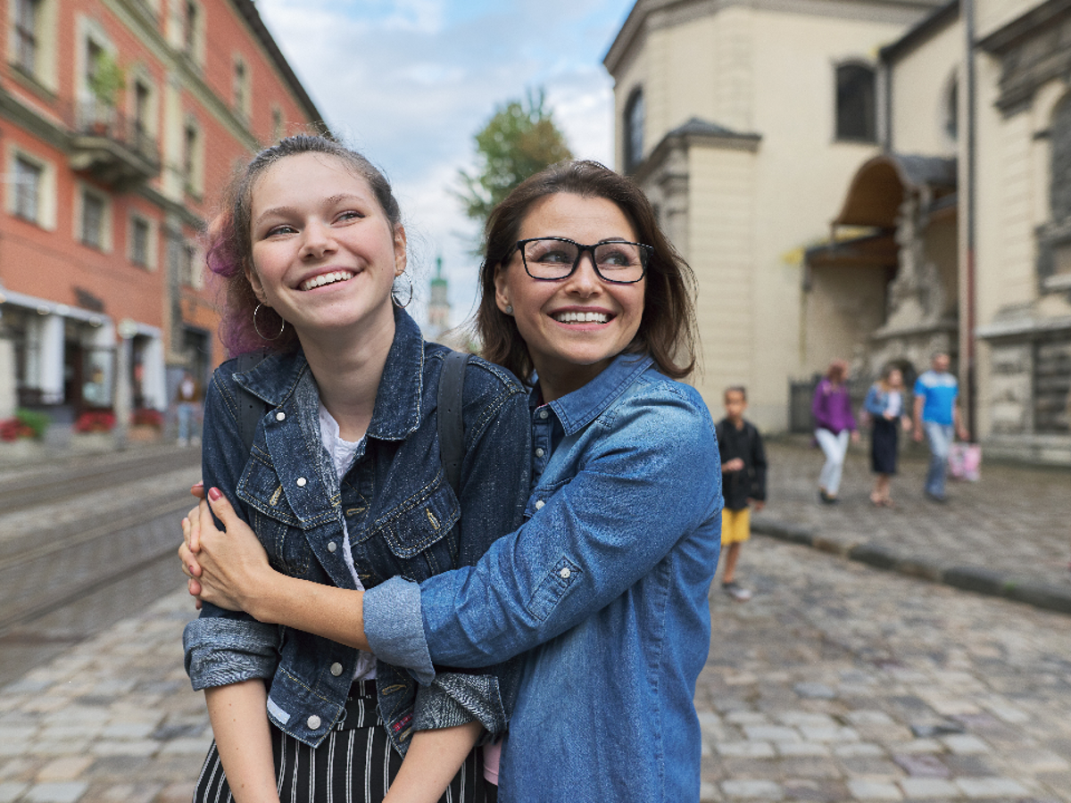 Two teenage girls smiling whilst standing in the middle of a road. One of the girls is hugging the other girl.