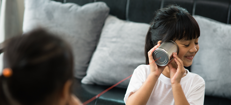 Two girls playing telephone with tin cans and string. Background is a grey sofa. Girl in foreground listening to tin can and smiling