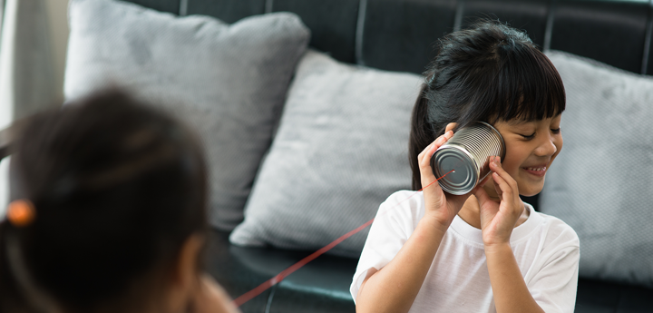 Two girls playing telephone with tin cans and string. Background is a grey sofa. Girl in foreground listening to tin can and smiling