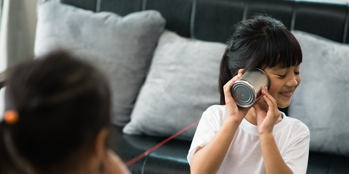 Two girls playing telephone with tin cans and string. Background is a grey sofa. Girl in foreground listening to tin can and smiling