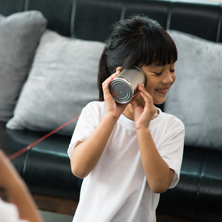 Two girls playing telephone with tin cans and string. Background is a grey sofa. Girl in foreground listening to tin can and smiling