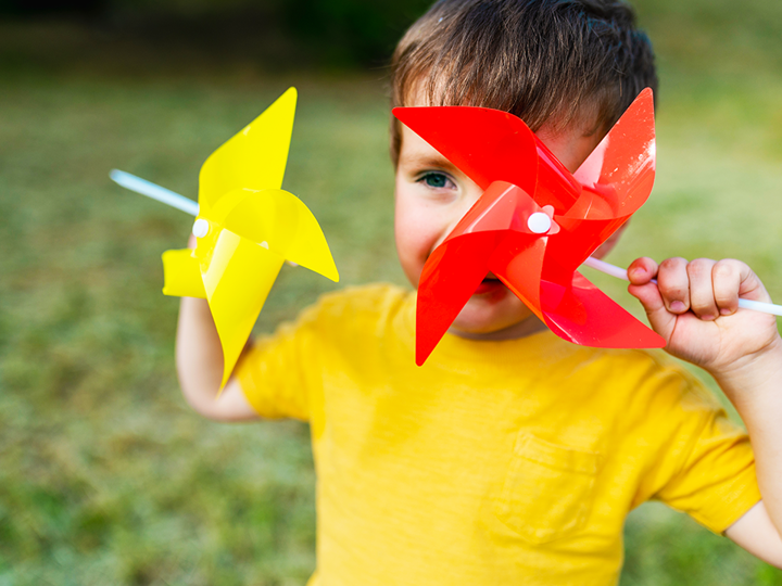 Boy holding 2 pinwheels to his face and smiling. 1 eye is visible through the pinwheels.