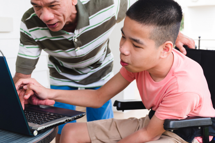 young man in wheelchair using a laptop with an older man leaning over to help