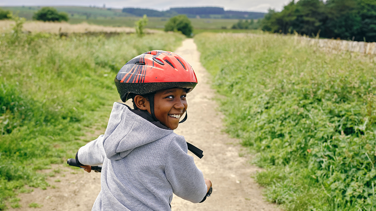 Child cycling on a dirt pathway in between 2 fields. They are wearing a red helmet and are looking over their shoulder