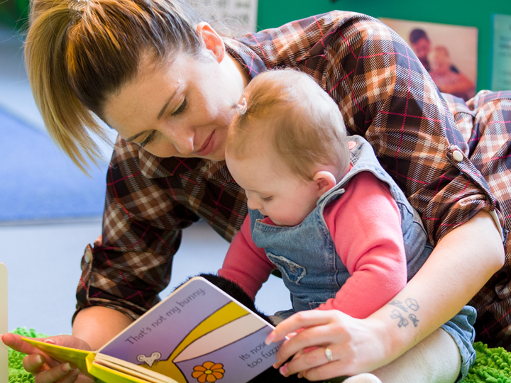 Adult leaning over baby girl who is sitting up and reading a book to her.