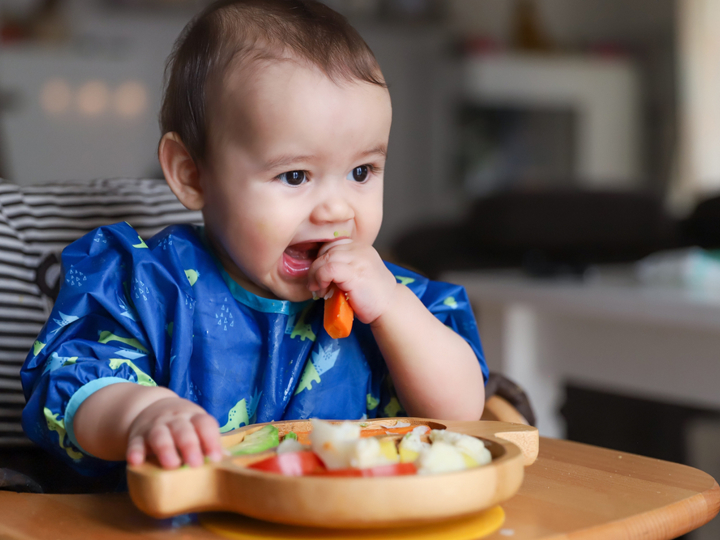 Baby boy sitting in highchair holding a carrot stick to his mouth with a plate of mixed vegetables in front of him.