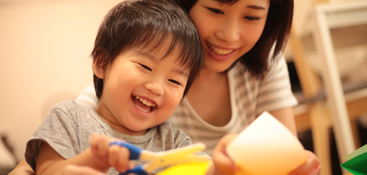 Child sitting on the lap of an adult. Parent is supporting their child cutting paper.