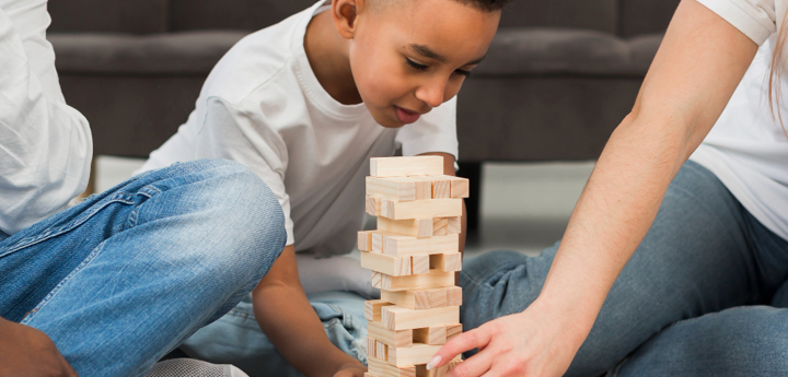 Young boy sitting on the floor playing Jenga next to 2 adults