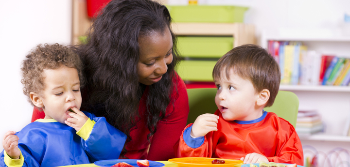 Two toddlers eating fruit sitting at a child's table in a nursery setting. A carer is sitting behind them. 